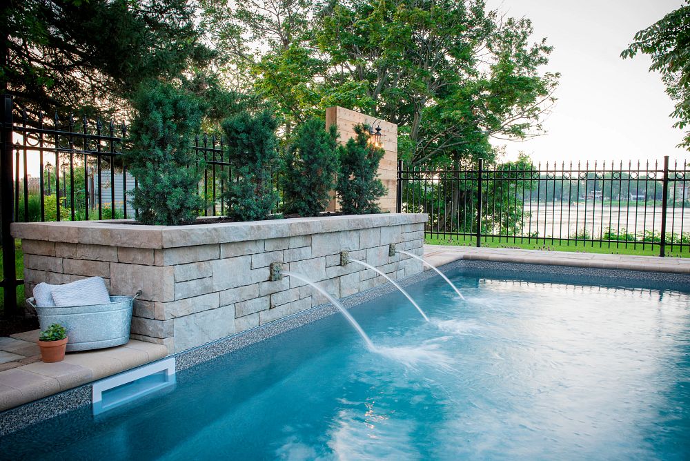 A luxurious backyard oasis featuring a pool with three fountains, enclosed by a gate, on a day with overcast skies.